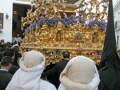 Un grupo de personas vestidas con capuchas blancas observan una procesión religiosa. La imagen muestra un altar dorado con adornos florales y figuras de oro, típico de una celebración religiosa. La escena se desarrolla en un entorno urbano, con edificios y personas alrededor.