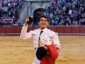Un torero en el centro de la imagen, con un traje blanco y rojo, sonríe mientras sostiene una muleta en la mano izquierda. La arena de toros está llena de espectadores detrás de una cerca roja.