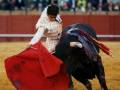 Un torero en traje tradicional sostiene un capote rojo mientras lucha con un toro negro en una plaza de toros. La arena está llena de espectadores observando la acción.
