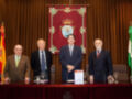 En una sala formal, cuatro hombres posan en un escenario frente a una bandera española y otra con un emblema. El hombre en el centro sostiene un libro, mientras los demás miran hacia la cámara. La decoración y el ambiente sugieren una ceremonia o conferencia oficial.