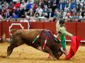 Un torero en traje verde y amarillo se enfrenta a un toro de color marrón en una plaza de toros. El torero sostiene una capa roja y se prepara para el combate. La arena es de color arena dorada, y la audiencia está atenta a la acción.