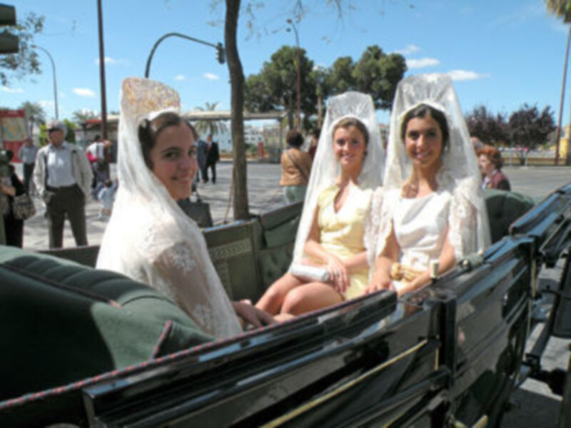 Tres mujeres vestidas con mantillas blancas y mantos de encaje se sientan en un carruaje de época, en un entorno urbano soleado.