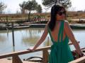 Elegante mujer en un vestido verde posando frente a un estanque con barcos y vegetación en el fondo.