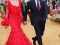 Una pareja camina por un ambiente festivo, la mujer viste un vestido rojo largo y el hombre un traje negro.