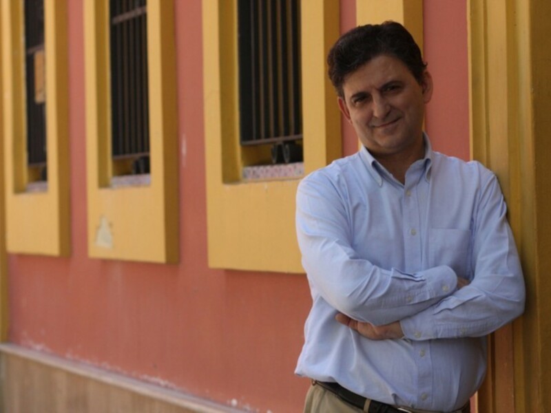 Un hombre con camisa azul y cinturón negro se apoya en una pared de color naranja y amarillo, frente a dos ventanas con persianas negras.