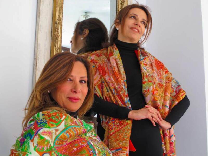 Dos mujeres posan con bufandas de colores vibrantes frente a un espejo. La escena parece ser en un ambiente elegante, posiblemente una boutique o estudio de moda.