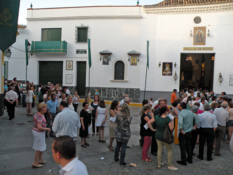 Una multitud en un evento público, frente a una casa blanca con detalles arquitectónicos. La imagen muestra a personas de diferentes edades y estilos, algunas con bolsos y otros con vestimentas informales. La casa tiene ventanas decorativas y un pórtico, con una inscripción que parece ser el nombre de la institución. La escena sugiere un ambiente festivo o ceremonial, con gente reunida en una calle o plaza.