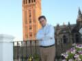 Un hombre posa con las manos cruzadas en un balcón con flores, con la Giralda de Sevilla en el fondo.