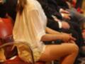 Una joven con un vestido blanco y sandalias rosa sentada en una silla de conferencia, rodeada por otros asistentes.