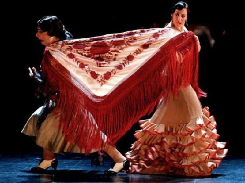 Danza flamenca con vestimenta tradicional y mantón de colores vibrantes en escenario iluminado.