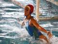 Una jugadora de waterpolo en acción, con un traje azul y casco rojo. La piscina está llena de agua transparente, con una red en el fondo y líneas de división. La jugadora está en movimiento, con las piernas extendidas y brazos levantados.