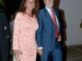 Una pareja en una reunión formal, el hombre lleva traje y corbata roja, la mujer usa un vestido corto con estampado floral en tonos de rosa y blanco.