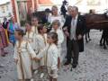Una boda tradicional en un pueblo con una joven pareja casada, acompañados por niñas vestidas de blanco y dorado. Un hombre mayor con corbata y traje negro los guía hacia un carro de caballos. Fondo con edificios coloridos y gente observando la ceremonia.