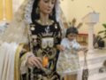 Imagen de una figura religiosa vestida con trajes tradicionales y un niño en sus brazos, posiblemente representando a la Virgen María. La escena se desarrolla en un ambiente religioso, con una base de madera y una alfombra roja.