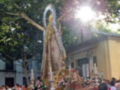 Una procesión religiosa con una estatua de la Virgen María en el centro, rodeada por flores y personas que parecen estar participando en la celebración. La estatua es dorada con detalles elaborados y tiene un halo de plumas. La escena se realiza al aire libre, con árboles y una estructura de madera en el fondo. La luz del sol brilla desde detrás, iluminando la escena y creando un ambiente festivo.