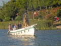 Barco blanco con una estatua en la parte delantera, flotando en el agua. Personas a bordo y alrededor. Fondo de vegetación y flores en el costado derecho.