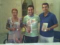 Tres personas sonrientes, dos hombres y una mujer, posando con tarjetas postales de diferentes lugares. La imagen parece estar en un entorno interior, posiblemente una sala o oficina, con una ventana y carteles en el fondo.