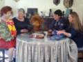 Una familia reunida en su hogar, con una mujer adulta y dos jóvenes sentadas a la mesa. La mesa está cubierta con un mantel de tela y sobre ella hay botellas, una figura de peluche, y otros objetos. La decoración del entorno sugiere un ambiente familiar y acogedor.