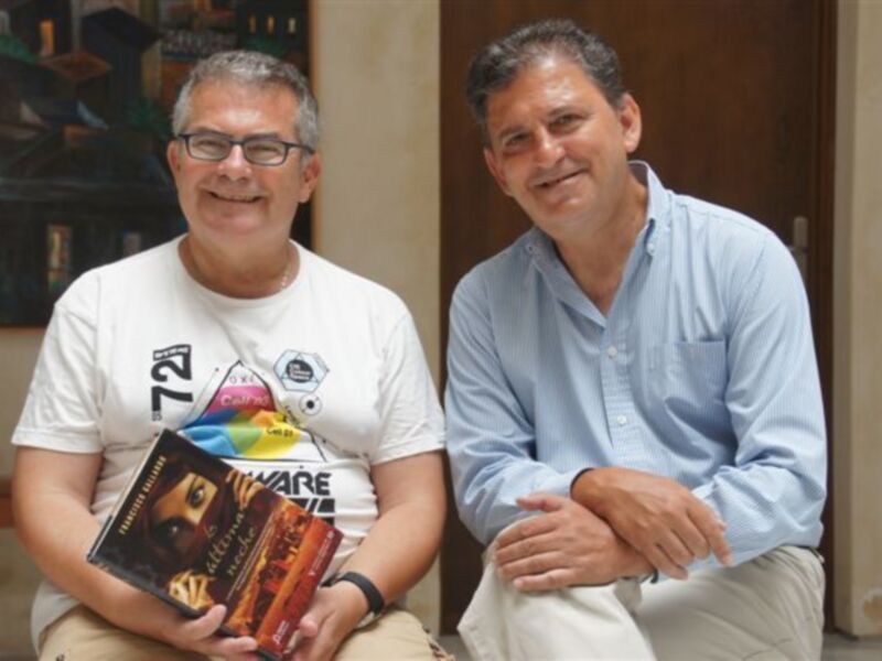 Dos hombres sonrientes posan con un libro titulado 'Alimentación y Nutrición' en un entorno acogedor.