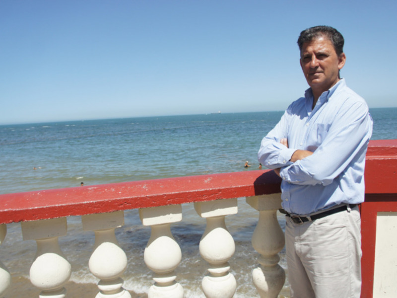 Hombre de pie en un muelle rojo frente al mar, con el cielo despejado y personas nadando al fondo.