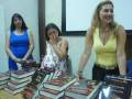 Tres mujeres sonríen mientras se colocan en una mesa llena de libros. La imagen parece ser parte de un evento literario o presentación de libros.