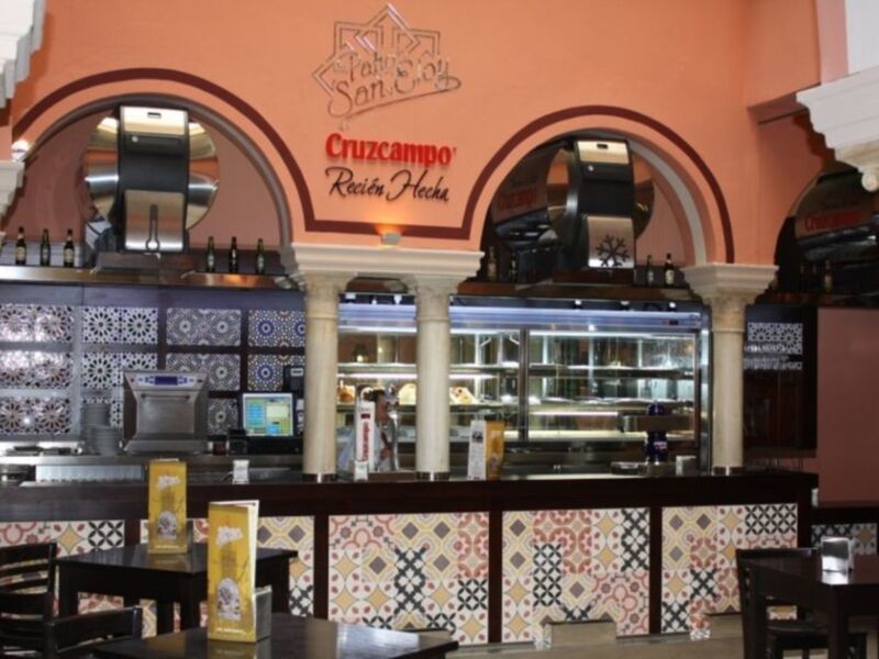 Interior de un restaurante con diseño tradicional, mostrando mesas, azulejos decorativos y una barra con marcas de cerveza Cruzcampo.