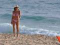 Una mujer en bikini rosa y sombrero de paja camina por la playa, con olas azules en el fondo.