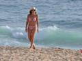 Una mujer en bikini rosa y sombrero blanco camina por la playa con olas azules detrás.