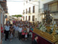 Una procesión religiosa en una calle de pueblo con gente vestida formalmente y un altar dorado con una estatua en el centro.
