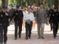 Una persona con una chaqueta blanca y pantalones negros es escoltada por varios oficiales de policía en un parque. La escena sugiere una situación formal o judicial, con la persona escoltada caminando entre los policías.