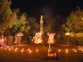 Una escena nocturna con tres personas vestidas de trajes tradicionales, posiblemente durante una celebración cultural. En el fondo, hay un monumento con una estatua y árboles iluminados. La escena está rodeada de farolillos que crean un ambiente festivo y mágico.