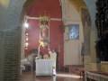 Interior de una iglesia con arcos y muros rojos. En el fondo, un altar con una figura en blanco y detalles dorados. La imagen muestra elementos arquitectónicos típicos de iglesias medievales.