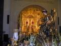Una escena religiosa en un altar dorado, con estatuas y flores blancas. La inscripción superior menciona a San Cristóbal de la Vera Cruz y Mina, y se observa una figura en trono con un sombrero.