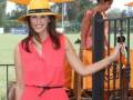 Una mujer con un vestido naranja y sombrero amarillo sonríe mientras se sube a una carruaje de jardín. La imagen muestra un ambiente festivo con otros asistentes en el fondo, y la luz del sol ilumina el entorno.