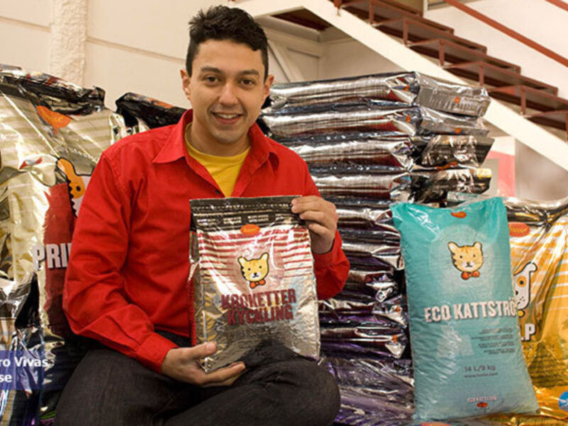 Un hombre sostiene una bolsa de comida para gatos en una tienda de mascotas, con varias bolsas de comida para gatos al fondo.