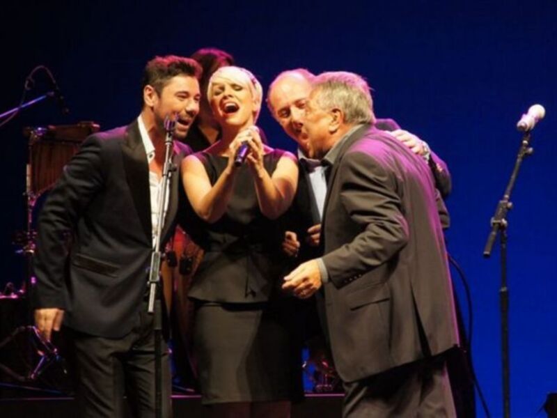 Tres personas en un escenario, dos hombres y una mujer, cantando o hablando. La mujer sostiene un micrófono y los hombres están abrazándola. El hombre a la izquierda lleva un traje negro y tiene una guitarra detrás de él. El hombre a la derecha también lleva un traje negro y está abrazando a la mujer. La escena es en una sala de conciertos con un fondo azul oscuro y luces brillantes.