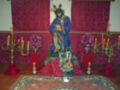 Estatua de Cristo en un altar con velas y flores, decorado con telas rojas y un tapiz ornamental en el fondo.