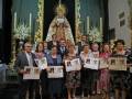 Una gran imagen religiosa con una Virgen en el centro, rodeada de un grupo de personas vestidas formalmente. Las mujeres están sosteniendo carteles con fotos y nombres, posiblemente en un evento religioso o festivo. La decoración incluye flores y velas, creando un ambiente festivo y reverente.