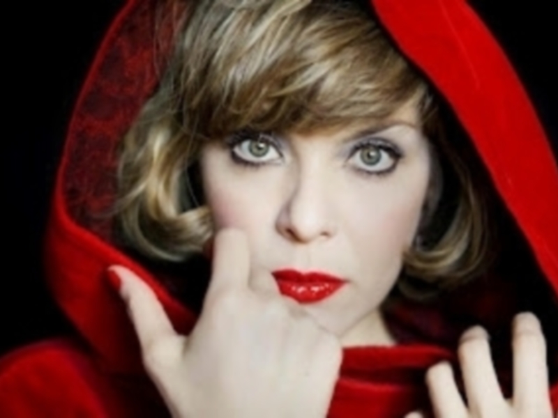 Una imagen de una mujer con una capucha roja y un maquillaje brillante, posando con una mano en la mejilla.