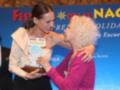 Dos mujeres en un evento de festivales, una entrega un premio a otra. Fondo con letras que dicen "Festivales de las Naciones".