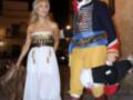 Una pareja vestida con trajes históricos, una en blanco y dorado y el otro en azul y rojo, caminando por la noche.