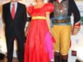 Tres personas posando en un evento. La persona de la izquierda está vestida con un traje negro formal, la central lleva una falda roja y rosa con detalles amarillos, mientras que la derecha está disfrazada de pirata. El fondo muestra carteles publicitarios con logos y nombres de empresas.