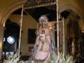Una procesión religiosa con una estatua de la Virgen María en el centro, rodeada de flores blancas y decoraciones. La escena se realiza dentro de una iglesia con arcos y vitrales, iluminada por luces que dan un ambiente sereno.