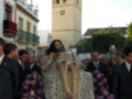 Una procesión religiosa en una ciudad española, con una figura vestida de blanco y rodeada por flores. En el fondo, se ve una torre con ventanas altas y un edificio histórico. La imagen transmite solemnidad y tradición.
