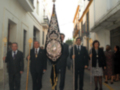 Una procesión religiosa en una calle de la ciudad, con personas vestidas formalmente y un personaje central con un traje ceremonial y un escudo en la espalda.