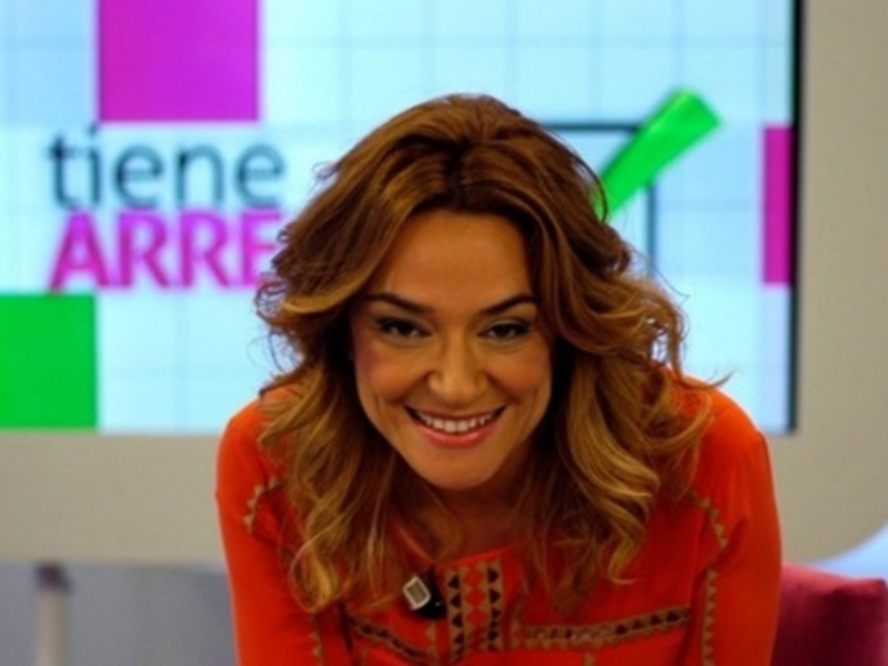 Presentadora sonriente en un estudio de televisión con fondo de pantalla en colores vibrantes.