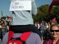 Una persona con una mochila roja sostiene a un niño que lleva un cartel que dice "No recorteis mi futuro". La imagen muestra a varias personas con carteles y banderas en un ambiente de protesta.