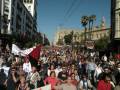 Una multitud de personas se manifiesta en una calle con pancartas y banderas, frente a un edificio histórico.