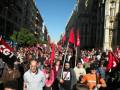 Una multitud en una manifestación, con banderas rojas y negras ondeando. La gente lleva camisetas con símbolos políticos, frente a edificios históricos. El cielo es azul claro y el ambiente parece ser de protesta o manifestación política.