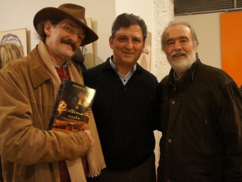 Tres hombres posan juntos en una galería de arte, uno sostiene un libro titulado 'La última noche'.